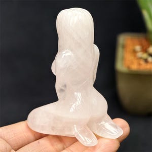 May include: A carved rose quartz mermaid figurine. The sculpture depicts a mermaid seated with her back to the viewer, her long hair flowing down her back. The figurine is a soft pink color and is held in a person's hand.