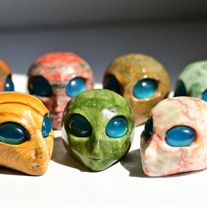 May include: A collection of alien head sculptures in various colors, including orange, red, green, and beige. Each head features large, blue, oval-shaped eyes. The sculptures are made of stone or a similar material.