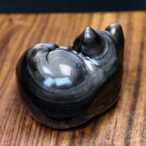 May include: A small, polished obsidian cat figurine in a curled-up position. The sculpture has a swirling pattern of black and gray, with a glossy finish. The cat's ears and tail are visible, and the figurine sits on a wooden surface.