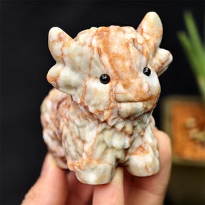 Puede incluir: Figura de piedra tallada de una vaca de las Highlands. La escultura es principalmente de color crema con vetas de color marrón rojizo. Tiene pequeños ojos negros y una expresión sonriente. La vaca está representada sentada.