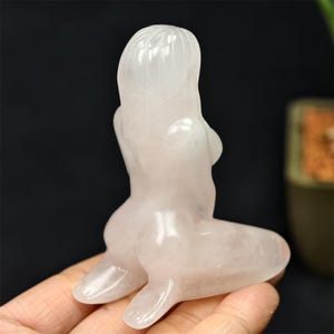 May include: A translucent, light pink crystal sculpture of a seated figure. The figure is kneeling with arms crossed, and long hair. The sculpture is smooth and polished, with a soft, glowing appearance. The figure is held in a hand.