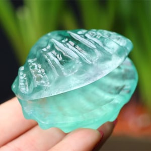 May include: A translucent, seafoam green seashell sculpture. The open shell reveals intricate details and textures. The sculpture is held in a hand, with a blurred green background.