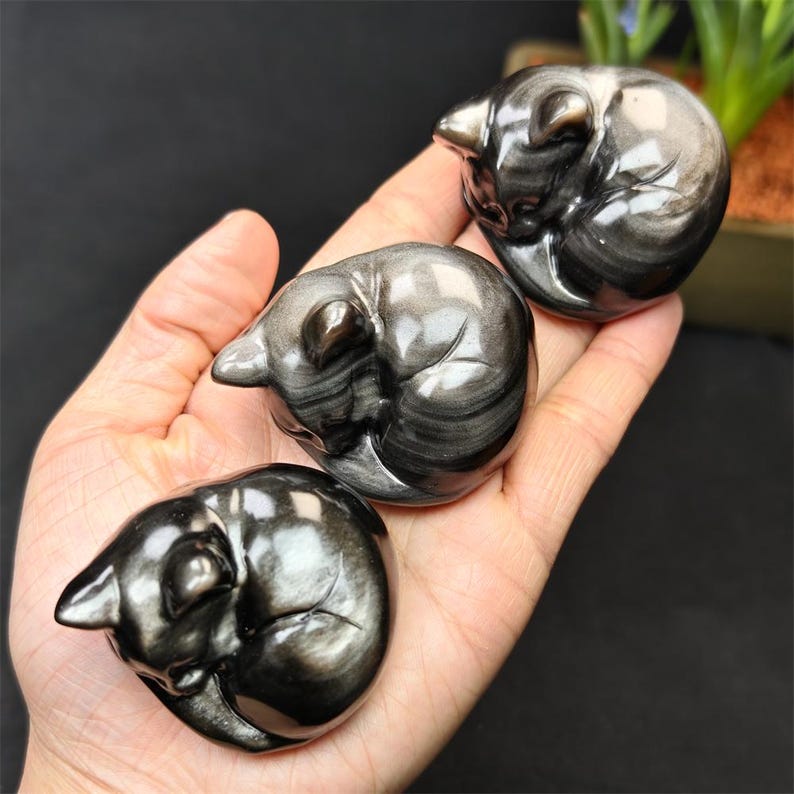 May include: Three polished obsidian cat figurines in a curled sleeping position. The cats have a metallic sheen with silver and black streaks. The figurines are held in a hand against a dark background.