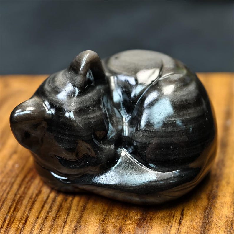 May include: A polished, black obsidian cat figurine in a curled-up sleeping position. The stone has a swirling pattern of gray and black, and the cat's form is smooth and rounded. The figurine is displayed on a wooden surface.