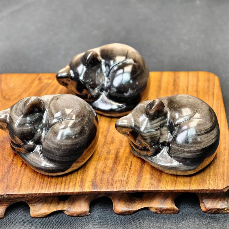 May include: Three polished obsidian bear figurines in a sleeping position, displayed on a wooden stand. The bears have a swirling pattern of black and gray, with a glossy finish. The wooden stand has a natural wood grain.