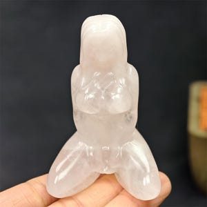 May include: A translucent, light pink crystal figurine of a kneeling person with long hair. The figure is carved with smooth lines, depicting the upper body and legs. The crystal has a polished finish.