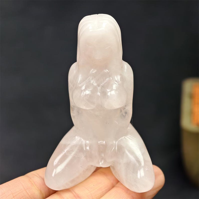 May include: A translucent, light pink crystal figurine of a kneeling person with long hair. The figure is carved with smooth lines, depicting the upper body and legs. The crystal has a polished finish.