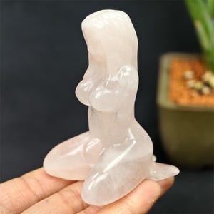 May include: A light pink, carved crystal figurine of a kneeling woman. The sculpture is smooth and polished, with details of the figure's form. The crystal has a translucent quality, and the overall aesthetic is delicate and artistic.