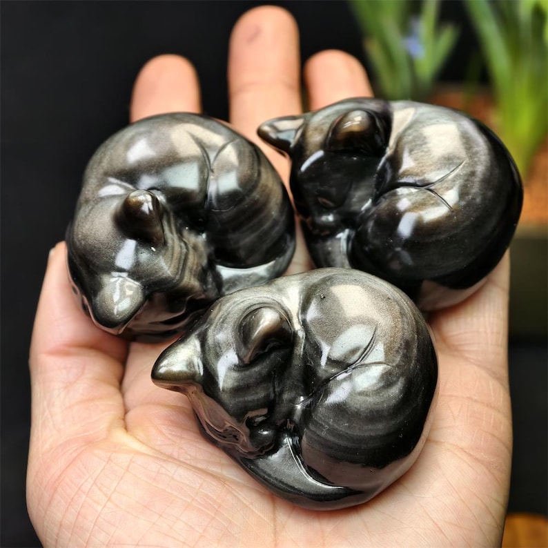 May include: Three polished obsidian cat figurines, each curled in a sleeping position. The cats have a reflective, metallic sheen with swirling patterns of black and silver. The figurines are held in a hand.