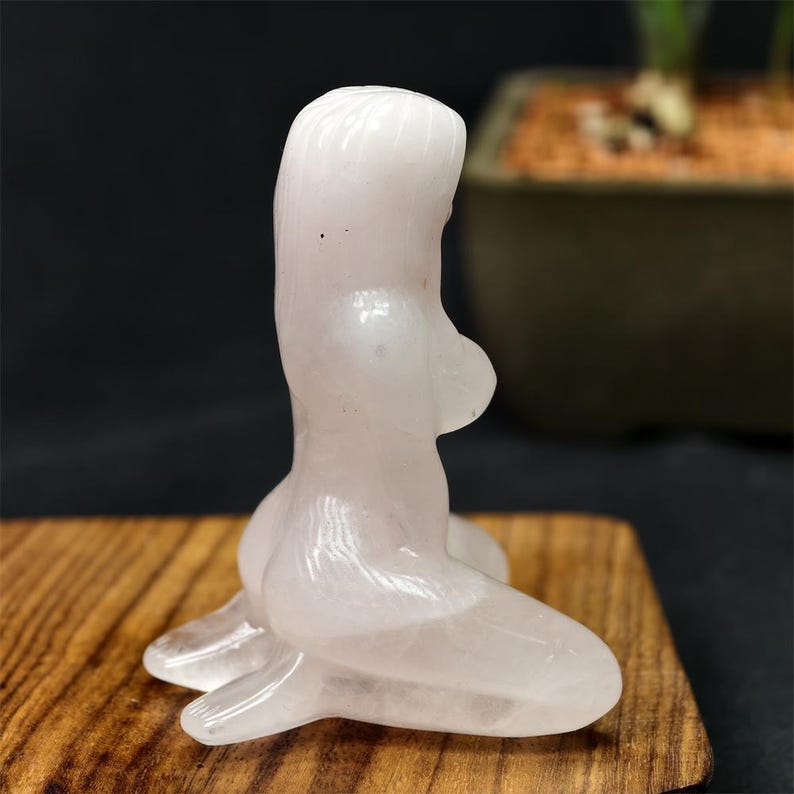 May include: A carved, light pink quartz figurine of a kneeling female form. The sculpture is smooth and polished, with rounded features and a subtle translucence. The figurine is displayed on a wooden surface.