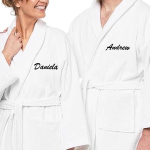 May include: Two white bathrobes with personalized embroidery. The robe on the left has the name "Daniela" embroidered on the left chest. The robe on the right has the name "Andrew" embroidered on the right chest.