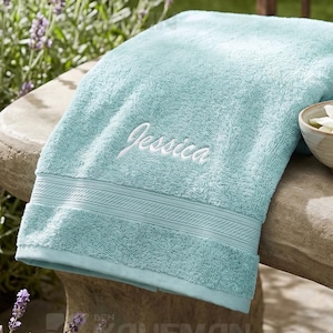 May include: A light blue bath towel with the name Jessica embroidered in white thread, featuring a textured border. Beside it sits a small rolled towel tied with twine, a ceramic bowl containing white and pink flowers, and a glass dropper bottle.