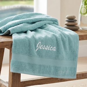 May include: A light teal bath towel featuring the name Jessica embroidered in white script lettering. The towel is folded on a wooden bench next to a clear glass carafe containing water and cucumber slices, a stack of smooth stones, and a white orchid.