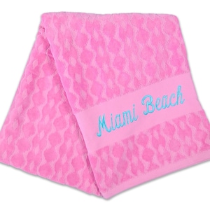 May include: A pink beach towel with a textured pattern and the embroidered text "Miami Beach" in blue. The towel is folded and displayed against a white background.