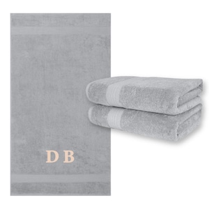May include: A light gray bath towel with the embroidered initials "DB" in a light pink color. Two folded matching towels are stacked to the right. The towels have a textured surface and a decorative band near the edge.