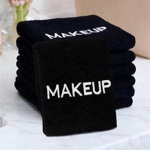 May include: A stack of black washcloths with the word "MAKEUP" embroidered in white. The washcloths are on a white countertop, with a makeup bag and a vase of pink flowers in the background.