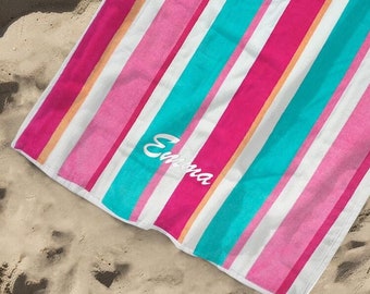 May include: A colorful beach towel with vertical stripes in pink, white, teal, and orange. The towel is lying on a sandy beach, with a straw hat and sunglasses nearby. The name "Emma" is written on the towel.