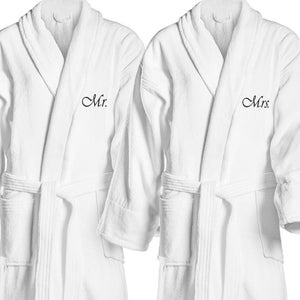 May include: Two white terry cloth bathrobes. One robe is embroidered with "Mr." in black script, and the other with "Mrs." Each robe has a shawl collar, a waist tie, and two front pockets. The robes are designed for comfort and relaxation.