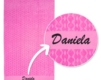 May include: A bright pink rectangular towel featuring a repeating geometric diamond pattern throughout the fabric. The towel includes a solid horizontal band near the bottom with the name Daniela embroidered in black script lettering.