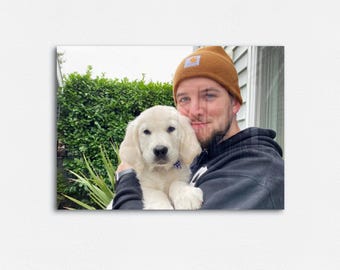 Puede incluir: Un cachorro joven de golden retriever con pelaje color crema es sostenido por una persona que lleva un gorro marrón y una sudadera con capucha negra. El cachorro tiene un collar azul. El fondo incluye follaje verde y una ventana.