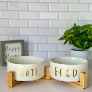May include: Two white ceramic pet bowls with gold lettering, one labeled "WATER" and the other "FOOD", are mounted on a bamboo stand.