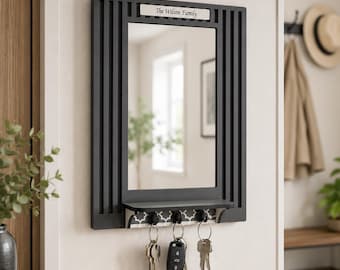 Puede incluir: Un espejo de pared rectangular negro con listones verticales en los laterales y un pequeño estante en la parte inferior. Una placa blanca en la parte superior muestra el texto The Wilson Family. Tres ganchos negros sostienen llaves de metal.