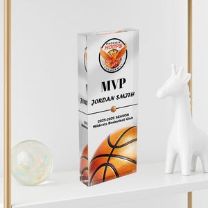 May include: A clear acrylic MVP trophy for the Phoenix Hoops Academy. The trophy features a basketball design and the text "MVP JORDAN SMITH 2025-2026 SEASON Wildcats Basketball Club". The trophy is displayed on a white shelf.