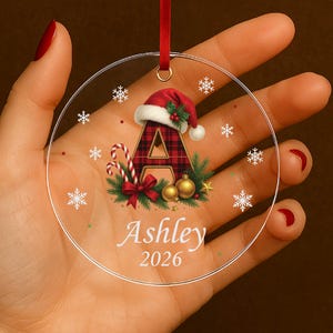 Pode incluir: Uma decoração de Natal redonda e transparente com uma fita vermelha. O enfeite apresenta a letra "A" xadrez com um gorro de Papai Noel, um bastão de doces, o nome "Ashley" e o ano "2026". Flocos de neve estão espalhados.