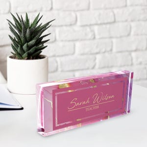May include: Pink acrylic desk nameplate with gold accents and the text "Sarah Wilson TEACHER" in a white font.
