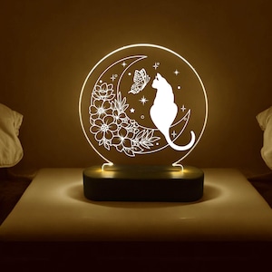 Könnte beinhalten: Eine weiße Katze sitzt auf einem Halbmond mit einem Schmetterling und einem Blumendesign. Das Design wird von einem Licht hinter der Acrylscheibe beleuchtet. Die Lampe steht auf einem Holzsockel.