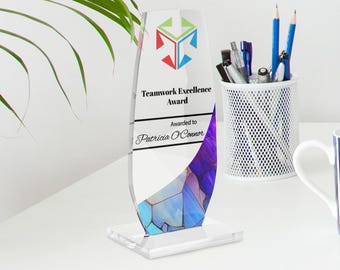 May include: A clear acrylic Teamwork Excellence Award with a colorful geometric design. The award has a rectangular base and an elongated, curved body. Text on the award reads "Teamwork Excellence Award" and "Awarded to Patricia O'Connor."
