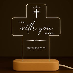 Può includere: Una luce notturna a LED a forma di croce con un contorno bianco. La croce presenta il testo "I AM with you ALWAYS" e "MATTHEW 28:20". È posizionata su una base di legno e ha un cavo di alimentazione.