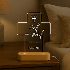 May include: A cross-shaped LED night light with the words "IT IS well WITH MY soul" and "PSALM 46:1" in white script. The light sits on a rectangular wooden base and is plugged into an electrical outlet.