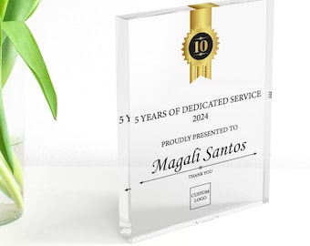 Puede incluir: Un premio de acrílico transparente con una cinta dorada y un sello negro y dorado de 10 años. El texto dice "5 Años de Servicio Dedicado 2024" y "Presentado con orgullo a Magali Santos". Incluye espacio para un logotipo personalizado.