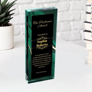 Puede incluir: Una placa de premio de vidrio verde y negro con letras doradas. La placa dice "Top Performer Award" y "Awarded to Sophia Roberts". El texto continúa con un mensaje de reconocimiento por el rendimiento excepcional y la búsqueda constante de la grandeza.