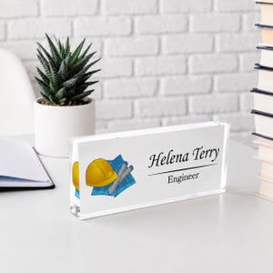 Puede incluir: Una placa de escritorio rectangular de acrílico transparente con el nombre impreso Helena Terry y el título Engineer. El diseño incluye un gráfico de un casco de seguridad amarillo sobre un plano arquitectónico azul con documentos enrollados.