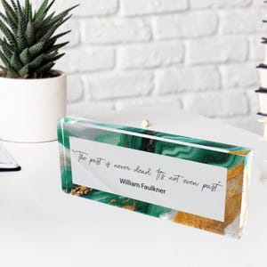 May include: A rectangular acrylic block with a green and gold marbled design. The block features a white rectangular insert with the quote "The past is never dead. It's not even past." by William Faulkner.