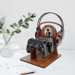 Può includere: Un supporto per giochi in legno con finitura marrone scuro, che contiene due controller di gioco neri e un paio di cuffie nere e rosse. Una foto circolare è centrata sul supporto. Il supporto è progettato per organizzare gli accessori di gioco.