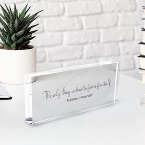 May include: Clear acrylic desk block with the quote "The only thing we have to fear is fear itself" by Franklin D. Roosevelt. The quote is in black script on a white background. A potted succulent plant is in the background.