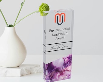 May include: A rectangular acrylic award with the text "Environmental Leadership Award" and the recipient's name. The award features a purple and white abstract design at the bottom and a logo at the top. A white vase with green flowers is in the background.