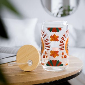 Könnte beinhalten: Klares, dosenförmiges Trinkglas mit Bambusdeckel und Strohhalm. Das Glas zeigt den Text "HAPPY TURKEY DAY!" in Orange und Weiß sowie herbstliche Blatt- und Fruchtdesigns. Der Deckel und der Strohhalm sind aus Bambus.