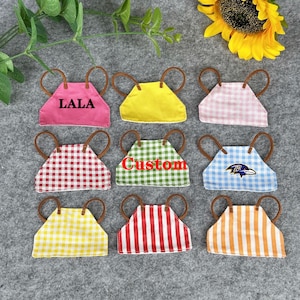 Könnte beinhalten: Neun Miniatur-Schürzen in verschiedenen Farben und Mustern, darunter Rosa, Gelb, rot-weißes Vichy-Karo und Blau mit Logo. Einige Schürzen tragen die Schriftzüge "LALA" und "Custom". Jede Schürze hat braune Griffe.
