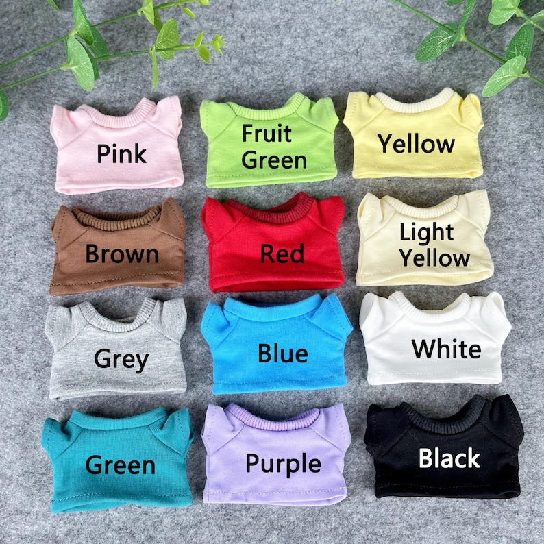 Pode incluir: Uma cole&ccedil;&atilde;o de nove camisetas em miniatura em v&aacute;rias cores, incluindo rosa, verde, amarelo, marrom, vermelho, amarelo claro, cinza, azul, branco, verde, roxo e preto. Cada camisa tem uma gola contrastante e o nome da cor impresso.