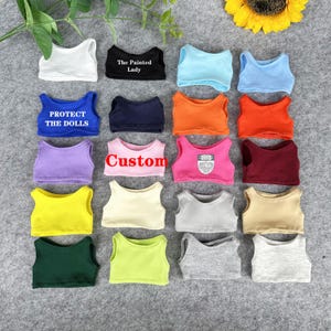 Peut inclure: Une collection de d&eacute;bardeurs pour poup&eacute;es de diff&eacute;rentes couleurs, dont blanc, noir, bleu, orange, rose, jaune, vert et gris. Certains hauts comportent des textes tels que "The Painted Lady", "Protect the Dolls" et "Custom".