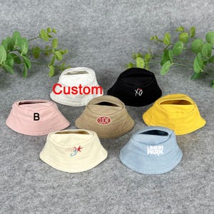 Könnte beinhalten: Eine Sammlung von sieben Fischerhüten in verschiedenen Farben, darunter Weiß, Schwarz, Rosa, Beige, Gelb und Hellblau. Jeder Hut hat einzigartige Texte oder Designs, wie "Custom", "XO", "B", "CLICHE" und "LINKIN PARK".