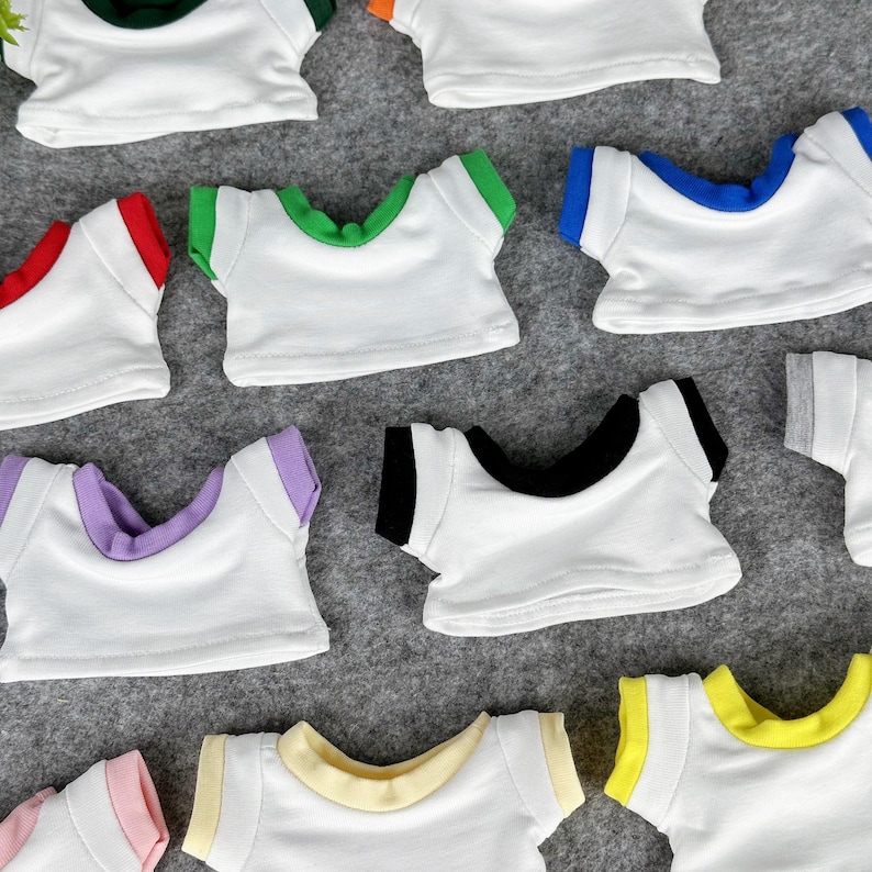 May include: A collection of small white t-shirts with colored trim around the neck and sleeves. The trim colors include red, green, blue, purple, black, yellow, and pink. The shirts are arranged on a gray surface.