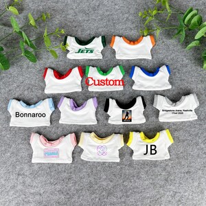 May include: A collection of small white t-shirts with various colored trim and text. The shirts feature text such as "Jets", "Custom", "Bonnaroo", and "JB". Some shirts have graphic designs.