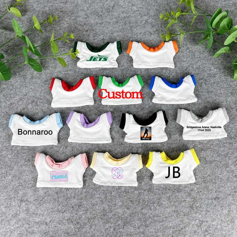 May include: A collection of small white t-shirts with various colored trim and text. The shirts feature text such as "Jets", "Custom", "Bonnaroo", and "JB". Some shirts have graphic designs.