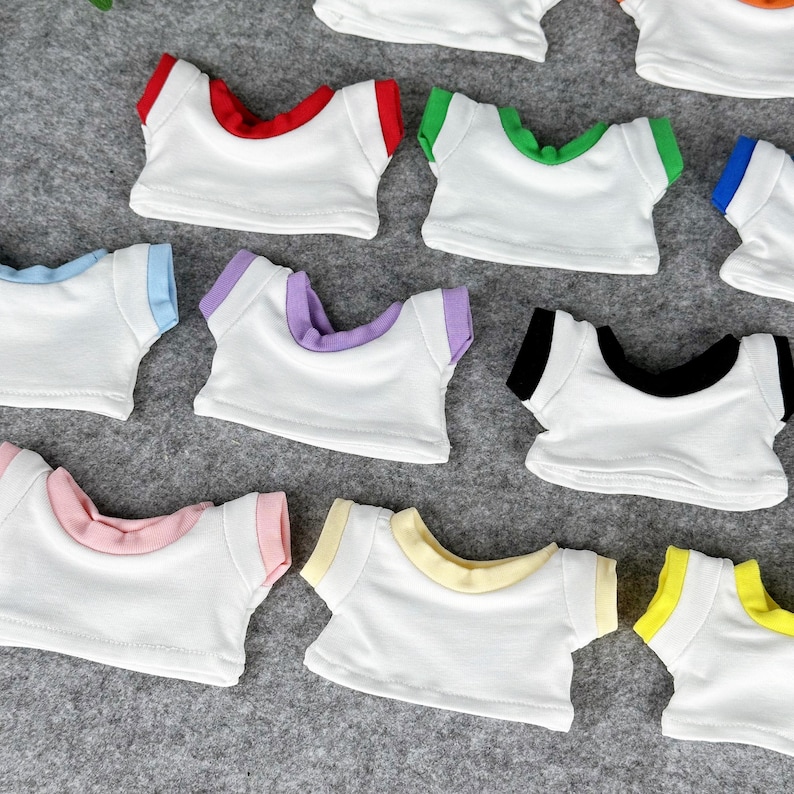 May include: A collection of small, white t-shirts with colorful trim in red, green, blue, purple, pink, yellow, and black. The shirts are arranged on a gray surface, showcasing the variety of color options.