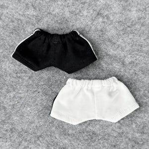 Puede incluir: Dos pares de pantalones cortos deportivos para mu&ntilde;ecas. Uno es negro con rayas laterales blancas, y el otro es blanco con rayas laterales negras. Ambos tienen cinturas el&aacute;sticas y est&aacute;n hechos de tela suave.